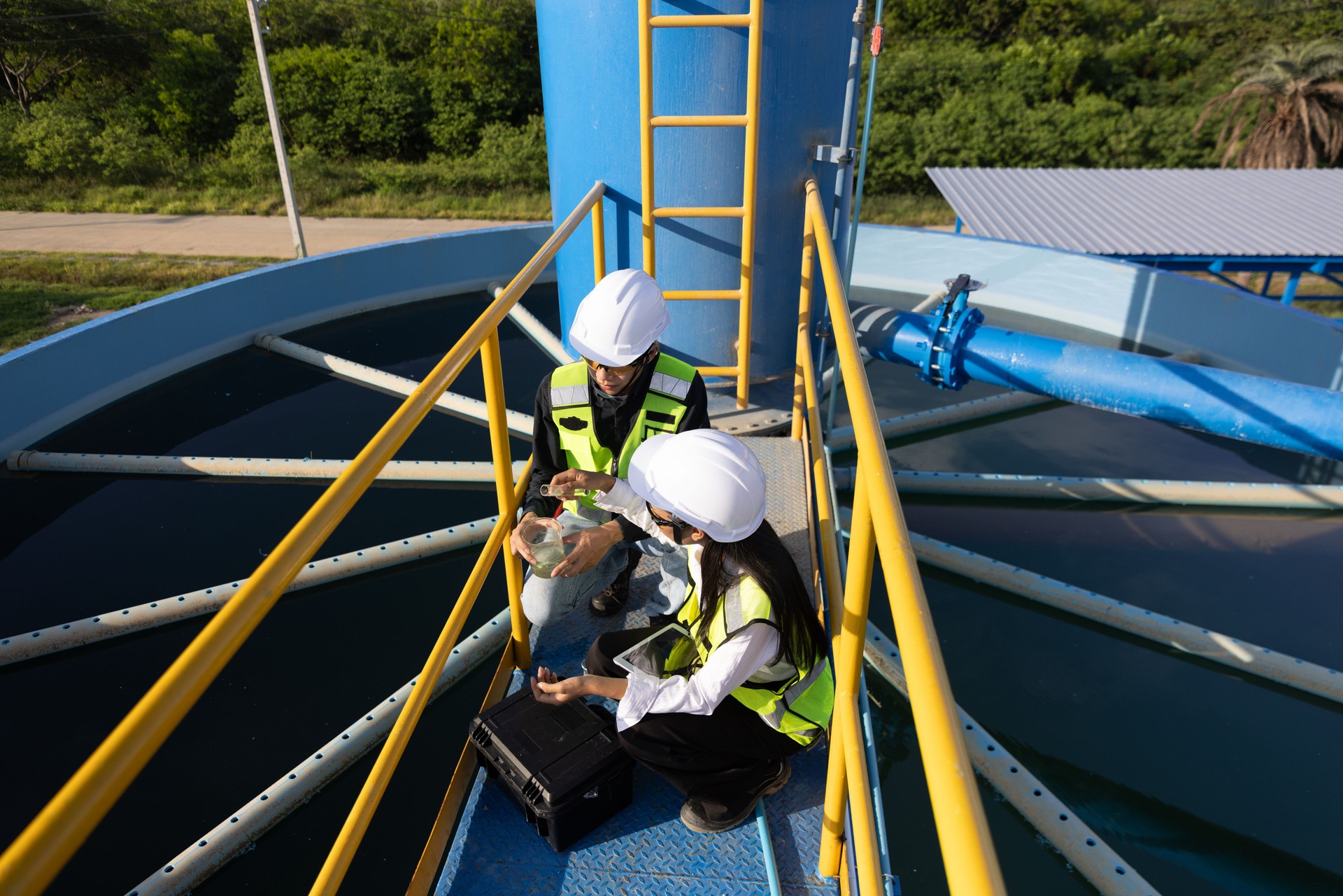 Two engineers wearing safety gear are inspecting and discussing documents on a clipboard while standing on a platform at an industrial water treatment facility, water management and environmental engineering